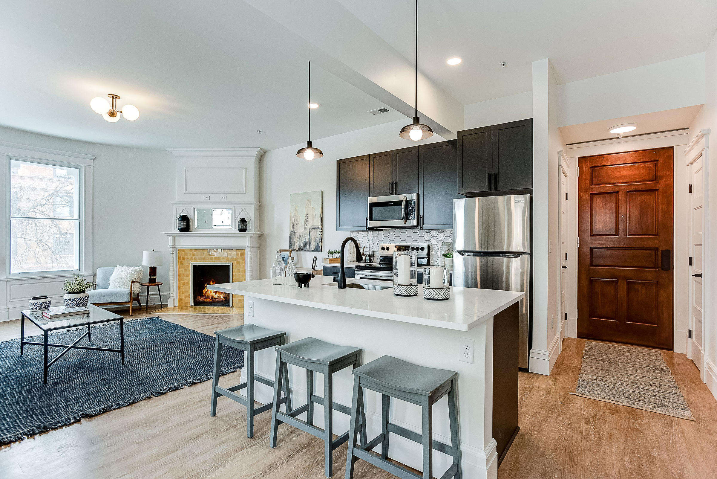 Modern kitchen with black cabinets, white island, and open living area with fireplace and large window.