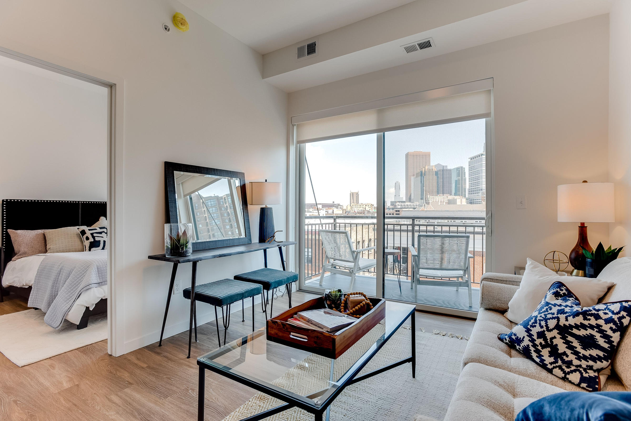 Modern living room with a sofa, glass coffee table, and balcony view of a cityscape; bedroom visible on left.