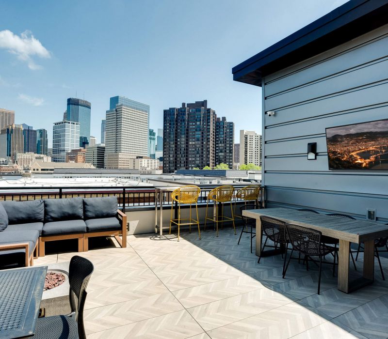 Rooftop patio with outdoor seating, a table, TV, and city skyline view under a clear blue sky.