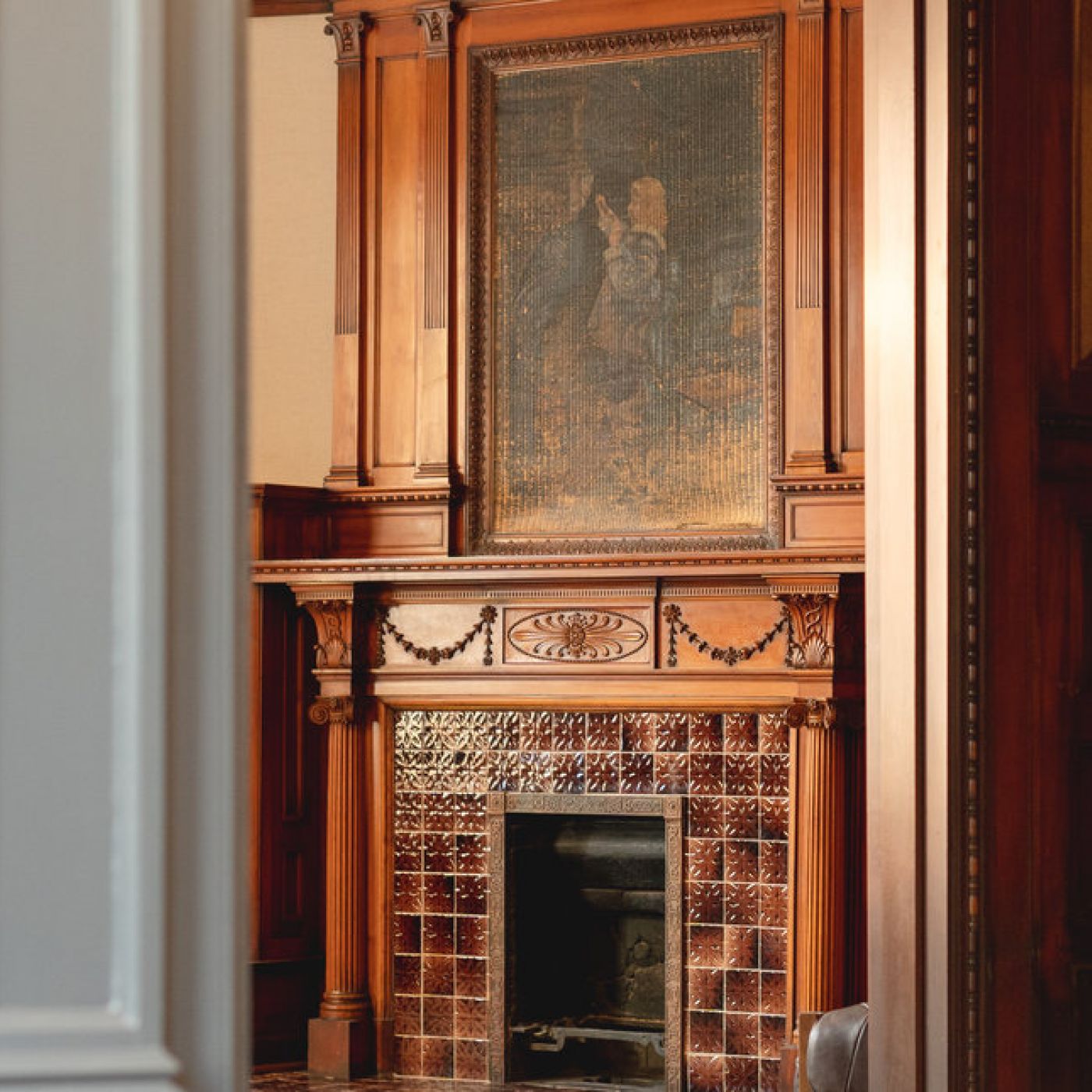 A wooden fireplace with decorative tiles and a large framed painting above it, viewed through a doorway.