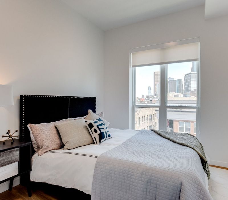 Modern bedroom with neatly made bed, nightstand, lamp, and large window showing a city skyline view.