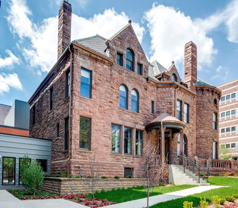 Historic stone building with arched windows and chimneys, surrounded by modern buildings under a blue sky.