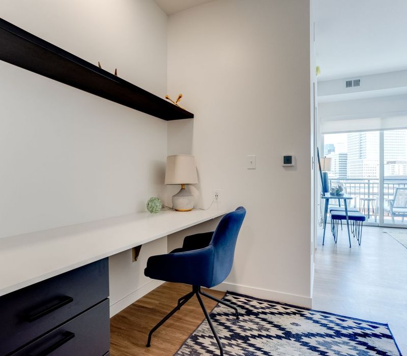 Modern home office with a blue chair, built-in desk, lamp, and view into a bright living area with cityscape.