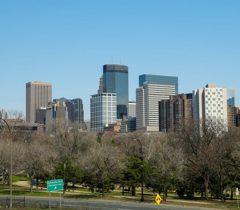 Downtown city skyline with tall buildings behind trees under a clear blue sky.