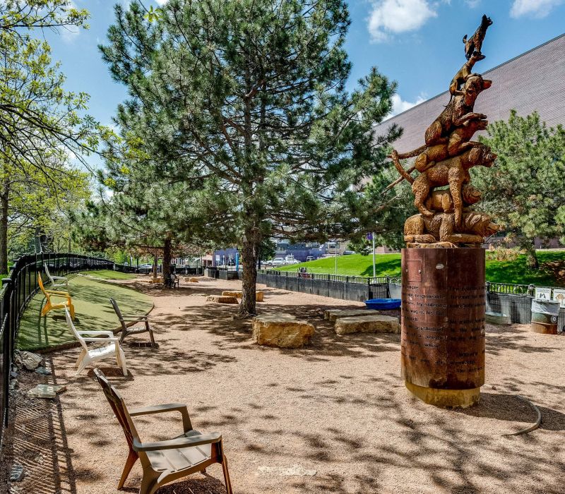 Fenced dog park with chairs, benches, trees, and a tall dog-themed sculpture on a sunny day.