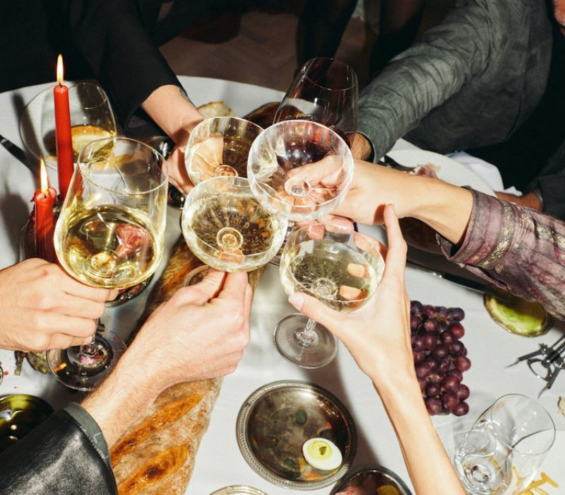 People clinking glasses of white wine over a festive dinner table with bread, grapes, and candles.