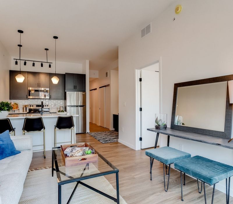 Modern apartment living room with sofa, glass coffee table, bar stools, and kitchen in the background.
