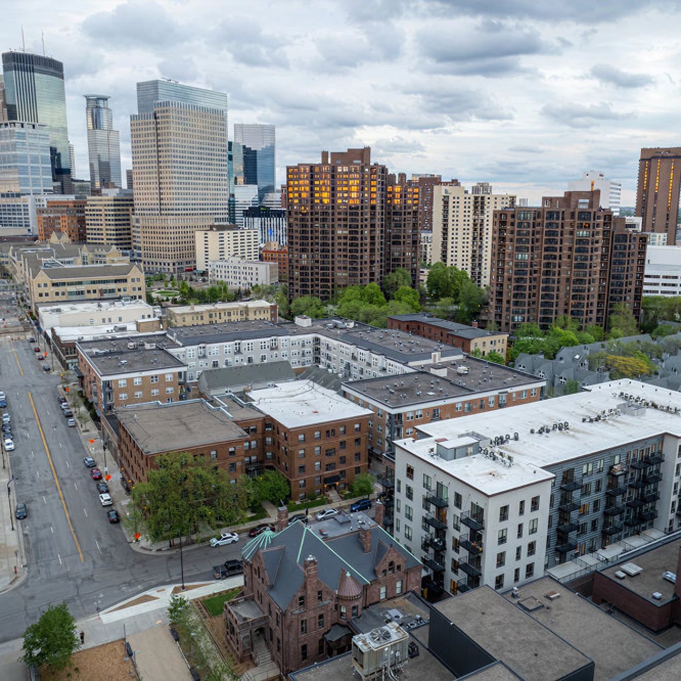 Aerial view of a cityscape with tall buildings, residential apartments, and tree-lined streets under a cloudy sky.