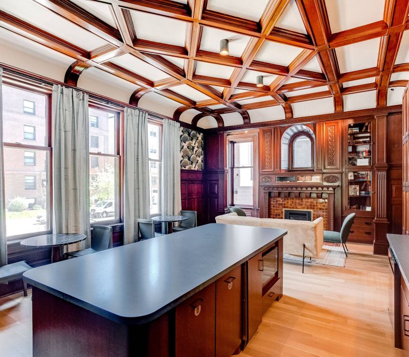 Elegant room with wood-paneled ceiling, island counter, seating area, fireplace, bookshelves, and large windows with curtains.