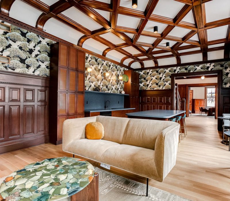 A stylish lounge with wood paneling, patterned wallpaper, a beige sofa, and a coffered ceiling.