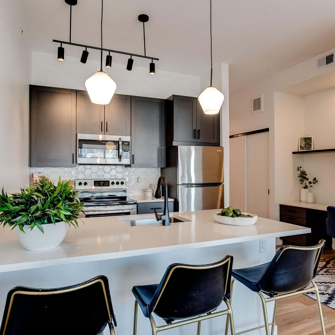 Modern kitchen with dark cabinets, stainless steel appliances, a white countertop, bar stools, and a small work nook with a desk and chair.