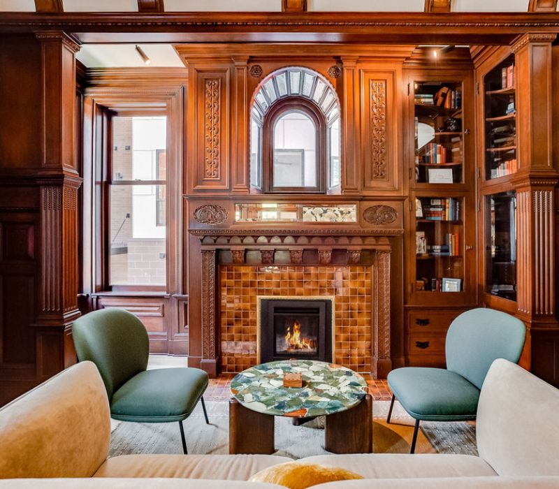 Elegant wood-paneled library with a fireplace, bookshelves, green chairs, and a marble coffee table.
