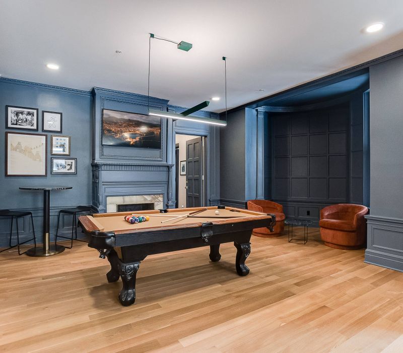 Modern game room with a pool table, wood floors, blue walls, fireplace, art, and two brown chairs.
